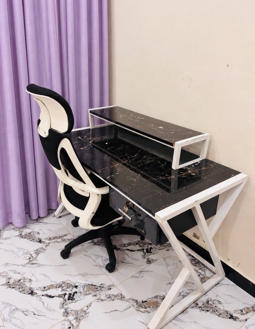 A modern computer desk with a black marble-patterned top and white Z-shaped legs, paired with a black and white ergonomic gaming chair, set against lavender curtains.