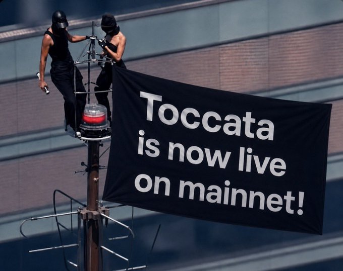 Two masked figures in black stand atop a tall pole, unfurling a large banner that reads "Toccata is now live on mainnet!" The dramatic, guerrilla-style display visually amplifies the breaking news announcement about Toccata's mainnet launch related to Kaspa ($KAS).
