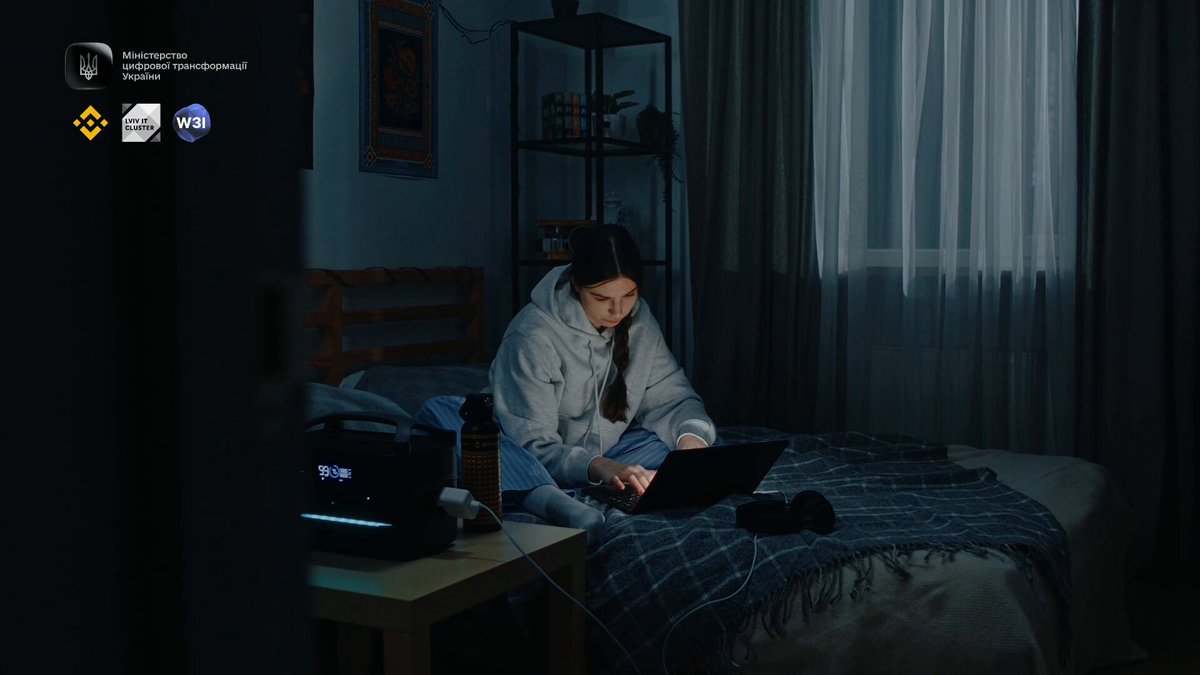 The image shows a woman sitting at her laptop, possibly working or browsing the internet. The room has a bed with a blue blanket and curtains, creating a cozy atmosphere. In the background, there are several logos, including one for Binance KYC that might be related to the image's context.