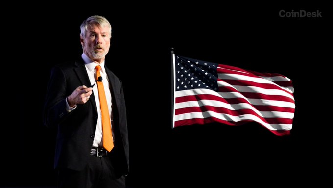 A man in a dark suit and orange tie stands at a podium gesturing with his hand, with a large waving American flag displayed behind him. The "CoinDesk" watermark appears in the upper right corner, and the image accompanies news about Michael Saylor and Strategy matching U.S. government contributions to Trump Accounts.