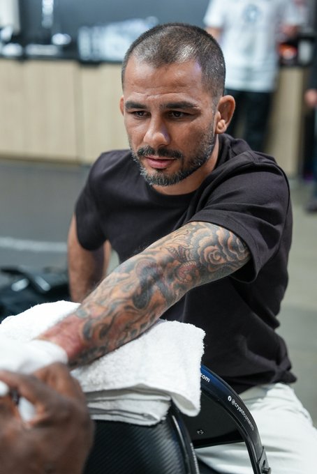 A focused MMA fighter with a closely-cropped haircut and intricate sleeve tattoo on his right arm sits in the corner, draped in a white towel over his shorts, displaying intense concentration between rounds. The image connects to the post by capturing the fighter's pre-fight or in-between-rounds mental preparation for the high-stakes CryptoCom-sponsored UFC 331 bout airing live on P+ Deportes.