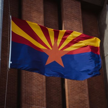 The Arizona state flag, featuring a red and yellow sunburst with a copper star over blue and red stripes, waves outside a brick building. The image relates to the post as it represents the state where Attorney General Kris Mayes has been actively combating crypto ATM fraud, helping victims recover significant funds.