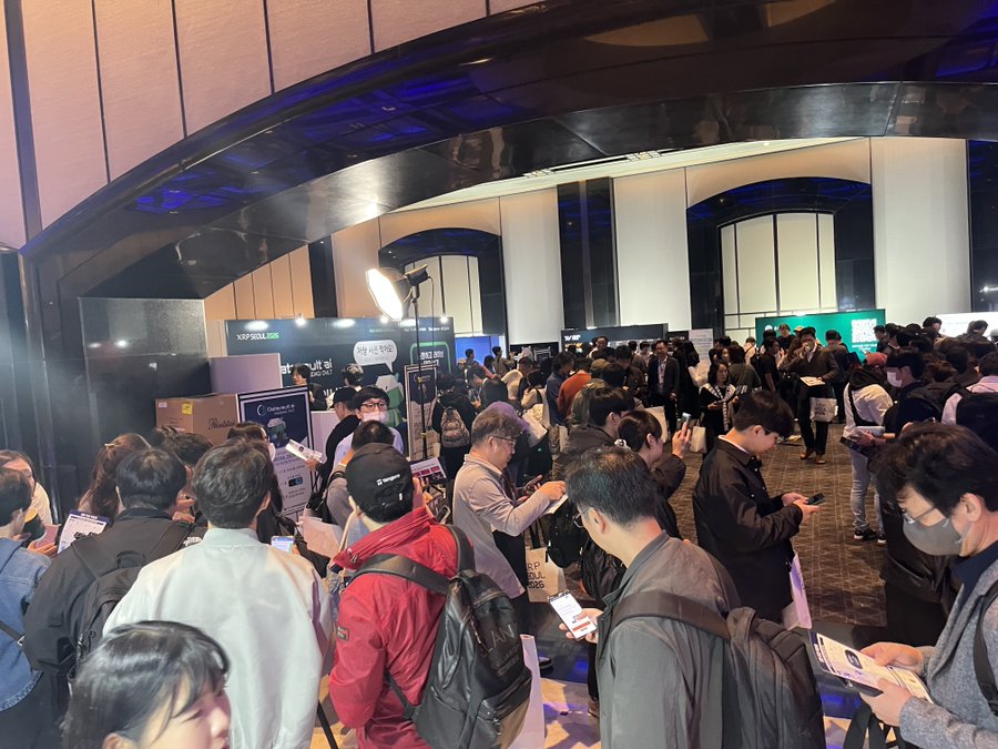 A crowded indoor crypto/Web3 conference with attendees at booths, including signs for 'XRP2POOL' and '해외코인', under dramatic blue lighting.