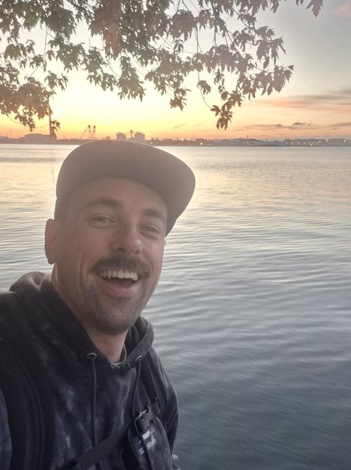 A man in a black cap and hoodie takes a selfie by a body of water during sunset, smiling at the camera with tree branches visible above.
