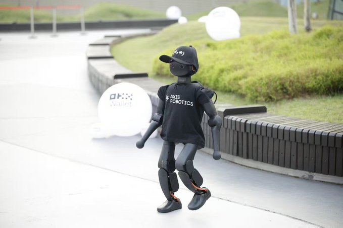 A humanoid robot wearing a black cap and an 'AXIS ROBOTICS' t-shirt walking along a paved path in a park, with white balloons in the background.