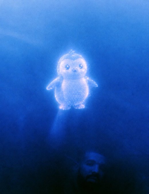 An underwater scene showing a glowing, sparkly blue penguin-like creature floating in the water. Below it, partially visible in the dark blue depths, is a man's face looking upward.