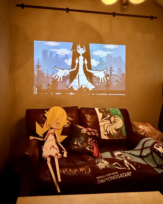 A room with a black leather couch displaying anime merchandise, including pillows with 'ONIMONOGATARI' branding and a cutout of a blonde anime character, while an anime character is projected onto the wall above.