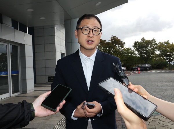 A man in a dark suit and glasses speaking to reporters, with multiple hands holding smartphones and recording devices extended toward him outside a building.