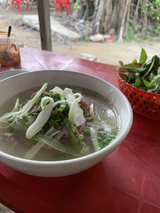 A bowl of noodle soup garnished with sliced white onions and green herbs sits on a red table next to a basket of fresh greens and a jar of red chili sauce.