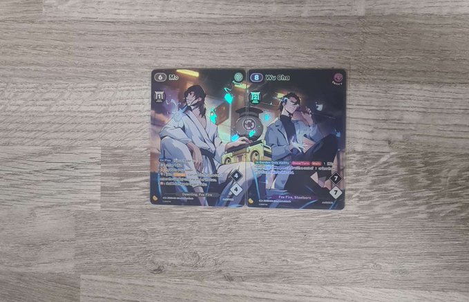 Two Azuki TCG character cards, "Me" and "Wu Sha," lie on a wooden surface, showcasing detailed anime-style artwork of a man in white and a woman in black. The cards' complementary designs visually connect, illustrating the post's point about the card art linking together, while the foil details highlight the set's premium finish.