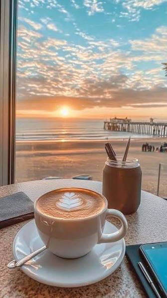 A latte with leaf latte art and an iced coffee sit on a table by a window overlooking a serene ocean sunset with a pier in the distance. The cozy seaside scene captures the perfect weekend moment to step away from the charts and enjoy quality time with loved ones.