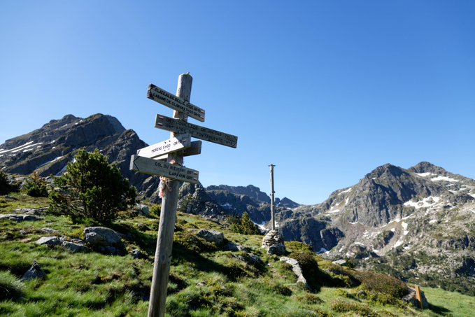 A wooden directional signpost with multiple arrows stands on a grassy alpine ridge under a clear blue sky, with rugged mountain peaks visible in the background.