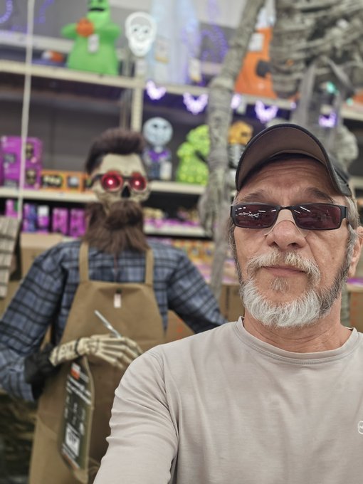 An older man with a gray goatee, wearing a black cap and red-tinted sunglasses, takes a selfie next to a Halloween animatronic skeleton wearing an apron in a Spirit Halloween store.