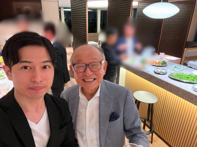 Two men, one older in a grey suit and glasses and one younger in a black jacket, pose for a selfie at what appears to be a restaurant or event with other people blurred in the background.