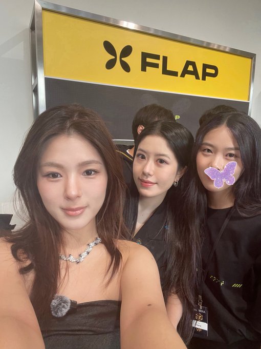 A selfie of three young women standing in front of a yellow sign with the word "FLAP" and a butterfly logo, with one woman on the right having a purple butterfly sticker covering her face.