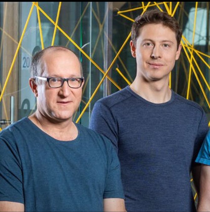 Two men stand side by side in blue shirts against a yellow geometric background.