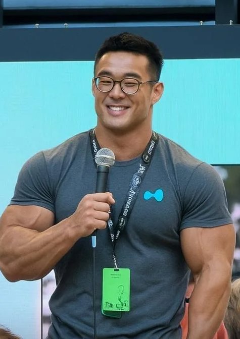 A smiling man wearing glasses and a gray t-shirt holds a microphone on a stage, with a green exhibitor badge around his neck.