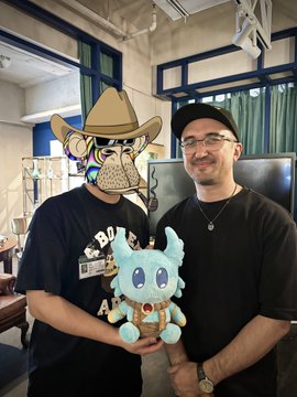 Two men pose together at a Bored Ape Yacht Club (BAYC) gathering, one wearing a hat with a cartoon ape graphic and holding a blue Bored Ape mascot figure, while the other smiles beside him—capturing the community-driven spirit highlighted in the post about Ape Toge and lasting connections formed through shared interest in the BAYC NFT collection.
