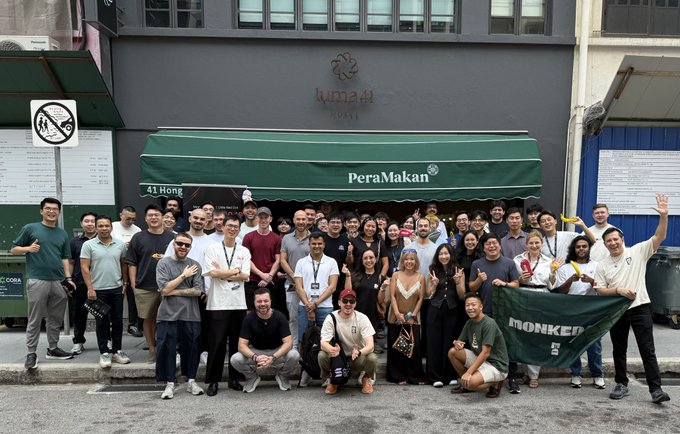 A large group of people posing for a photo in front of 'PeraMakan' restaurant at '41 Hong' and 'lumeH Hotel KL Sentral', with some holding a green 'MONKED' flag.