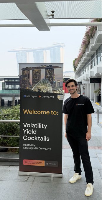 A man wearing a black Derive.xyz t-shirt stands next to a banner reading "Welcome to: Volatility Yield Cocktails," hosted by GTS Digital & Derive.xyz, with the iconic Marina Bay Sands visible in the background of Singapore. The image illustrates his first time hosting an event for Derive, capturing the celebratory setting of an onchain options gathering surrounded by Singapore's stunning skyline.