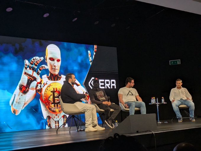A panel discussion on stage at what appears to be a crypto/blockchain event, with four men seated in chairs and a large screen behind them displaying a robot graphic holding a Bitcoin and the word 'ERA'.