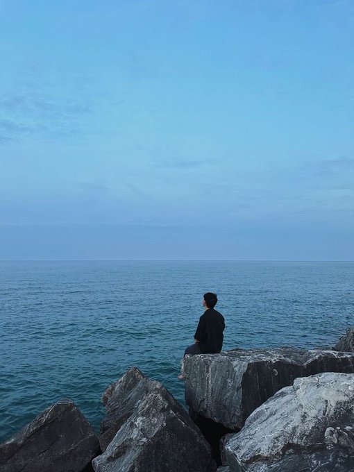 A serene photograph of a person dressed in dark clothing sitting alone on a large rock by the sea, gazing out over calm blue waters under a clear sky.