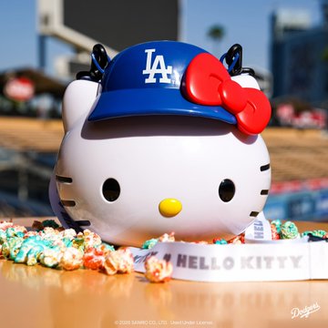 A Hello Kitty collectible figure wearing a blue Los Angeles Dodgers baseball cap with a red bow, sitting on popcorn at what appears to be Dodger Stadium, with 'HELLO KITTY' text on the base.
