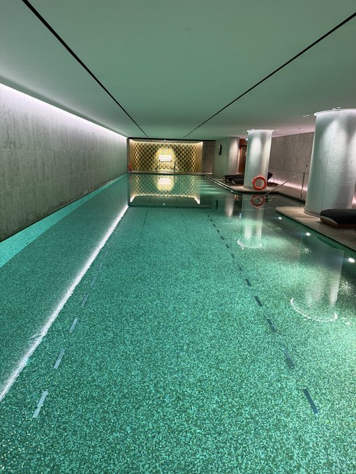 An empty, luxurious indoor swimming pool with teal mosaic tiles reflects a glowing golden doorway at the far end, flanked by columns and minimalist seating. The serene, unoccupied space mirrors the post's irony of a "jobless, purposeless crypto guy contributing absolutely nothing to society"—much like the lavish pool, it's beautiful, pristine, and going completely unused.