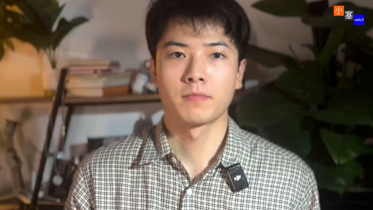 The image shows a man wearing a plaid shirt, with an electronic device attached to his shirt that has the word "predict" written on it. The man appears to be looking directly at the camera while posing for the photo. The background of the image features a potted plant and some bookshelves, suggesting that he might be in a room or office setting.