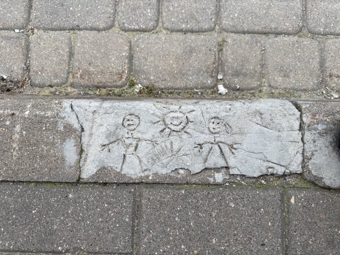 A stone curb on a paved sidewalk features a simple child-like carving of two stick figures with a smiling sun between them.