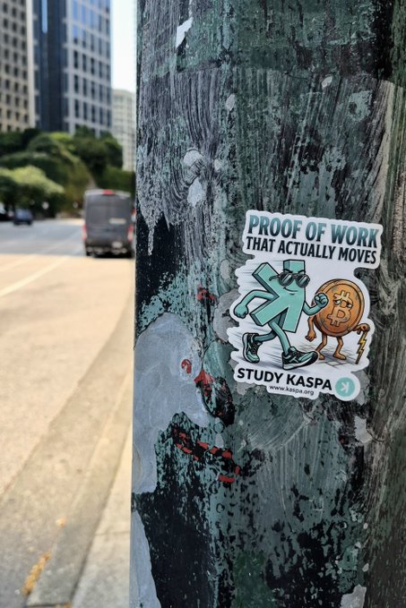 A sticker on a weathered urban pole reading 'PROOF OF WORK THAT ACTUALLY MOVES' with 'STUDY KASPA www.kaspa.org', depicting a figure carrying a Bitcoin coin, set against a city street backdrop.