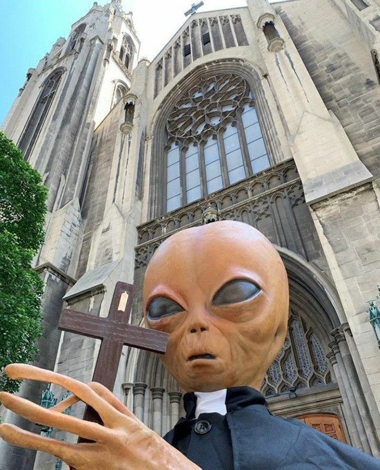 A person wearing an alien mask and holding a wooden cross stands in front of a large Gothic cathedral.