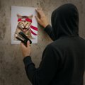 An anonymous figure in a dark hoodie is putting up a poster of a cartoon raccoon character on a concrete wall, referencing the $GME and @GMEethereum tags, likely symbolizing the playful, meme-driven "diamond hands" culture and community spirit within the GameStop and crypto investor movements.