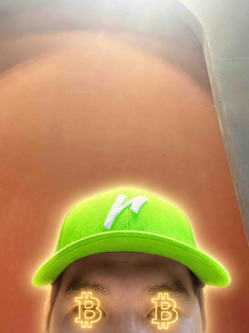 A close-up selfie of a person wearing a neon green cap with an 'n' logo, featuring glowing Bitcoin symbols edited over their eyes.