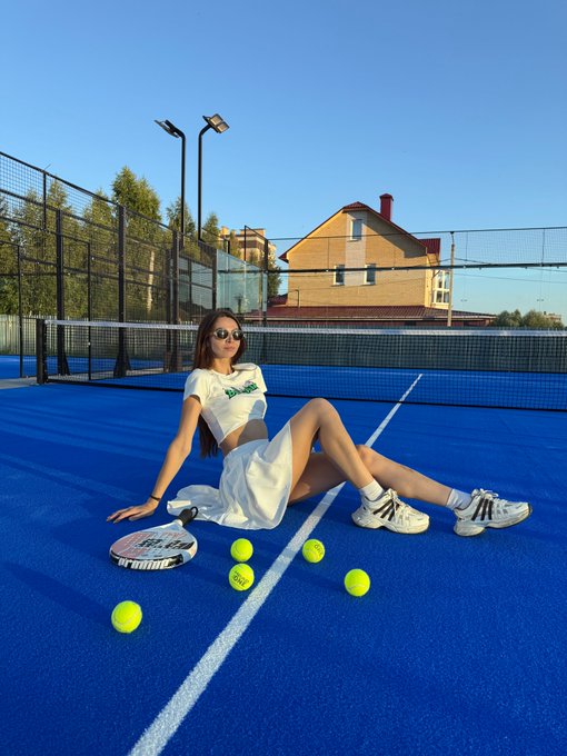 A young woman wearing sunglasses, a white 'Barbie' crop top, and a white skirt sitting on a blue tennis court, holding a racket, with yellow tennis balls scattered around her.