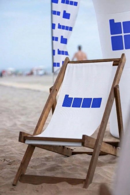 The image features a beach scene with a white beach chair positioned near the water's edge, facing away from the camera. The chair has blue letters on its backrest that say "base" and "#base #jessepollak @coinbase". In the background, there are people enjoying their time at the beach, as well as a few umbrellas providing shade for those who prefer to stay out of the sun.