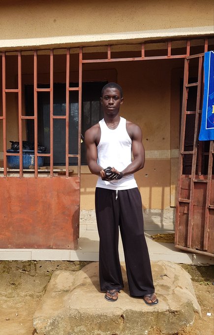 A young man wearing a white tank top and dark pants stands on a rock in front of a building with red barred windows and doors.