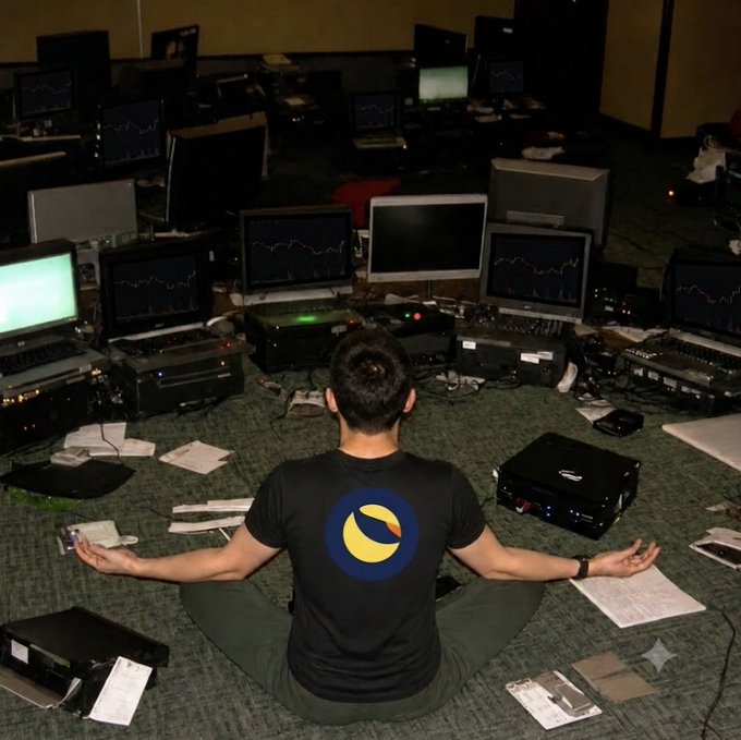 A person wearing a black t-shirt with a yellow and blue circular logo sits cross-legged in meditation pose on the floor of a room filled with multiple computer monitors displaying stock charts. The image evokes a sense of calm focus and readiness, symbolizing the community's meditative optimism and preparation for Terra Classic (LUNC) to make a comeback.