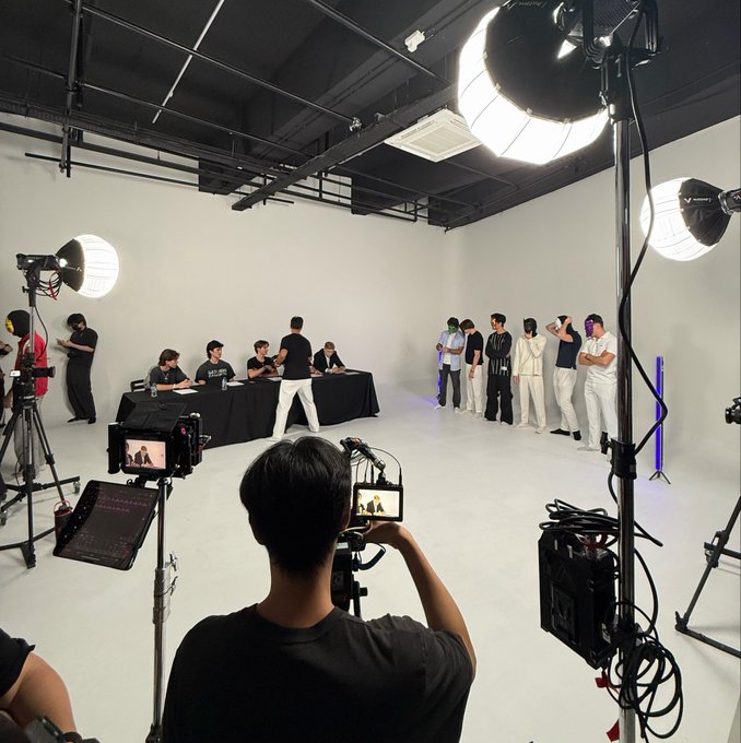 A behind-the-scenes view of a professional photo/video shoot in a white studio, showing crew members operating cameras and lighting equipment while a group of performers in white outfits stands in the background.
