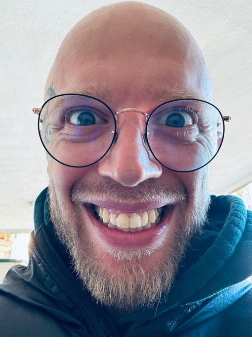 A close-up selfie shows a bald man with round glasses, a ginger beard, and an exaggerated wide-eyed, toothy grin, capturing the manic energy of someone chasing degen trades while dodging accountability. The caption leans into the post's plea for public tickers to blame for losses, with his over-the-top expression perfectly embodying the "degen without responsibility" spirit.