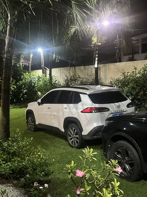 A nighttime photo of two SUVs parked on grass in a tropical setting, with palm trees and outdoor lights visible in the background.