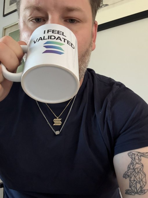 A man drinks from a white mug that reads "I FEEL VALIDATED" with a colorful logo, celebrating a "big week" with a joyful morning coffee moment. His dark navy t-shirt and geometric bear tattoo on his forearm add a personal touch to the triumphant snapshot.