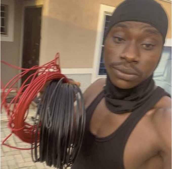 A selfie of a man wearing a black durag, black tank top, and a black neck gaiter pulled down, holding up a large bundle of tangled red and black electrical cables or wires.