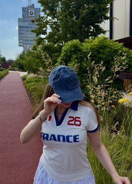 A casual outdoor photo of a young woman wearing a blue denim cap and a white 'France' sports jersey with the number 26, standing beside a garden path with a building in the background.