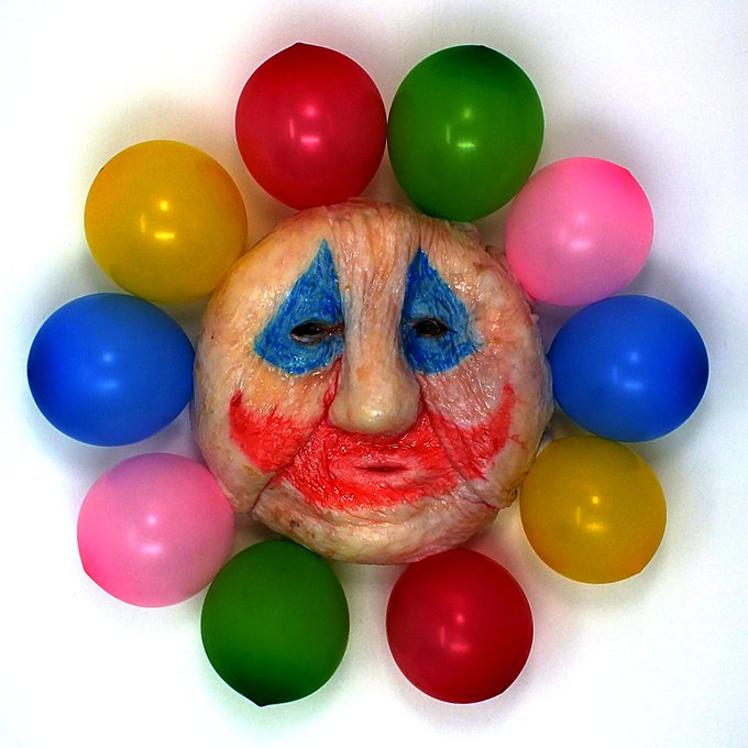 A grotesque, clown-like sculpted face with smeared blue and red makeup is surrounded by a ring of colorful balloons (red, green, pink, yellow, and blue), evoking the unsettling, childlike-yet-disturbing imagery associated with serial killer John Wayne Gacy, who was known for his "Pogo the Clown" persona—fittingly referenced in the "GACY (organic biological media)" caption.