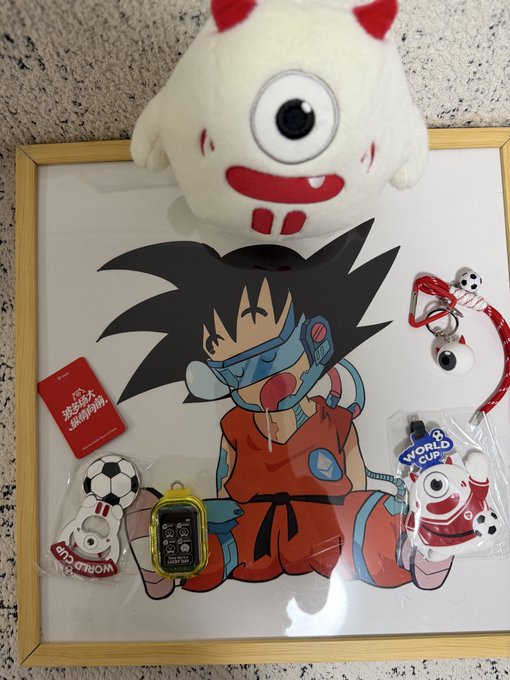 A framed illustration of a young Goku wearing a futuristic blue breathing apparatus is displayed alongside a "WORLD CUP" badge, a soccer ball mascot keychain, a white plush toy with horns, a small red card, and a handheld electronic device featuring an elephant design. The image connects to the post's theme of a "bullish" character collection (echoing the bull-like horns and red accents), with the elephant device likely representing the "metaphysics machine" used for fortune-telling.