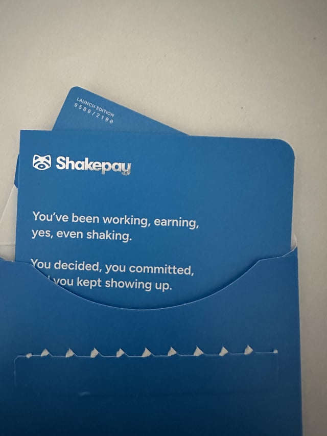 A blue Shakepay "Launch Edition" card (numbered 0660/2100) sits partially in its envelope, featuring the brand's tagline "You've been working, earning, yes, even shaking." The card visually corresponds to the post announcing the arrival of the new Shakepay card.