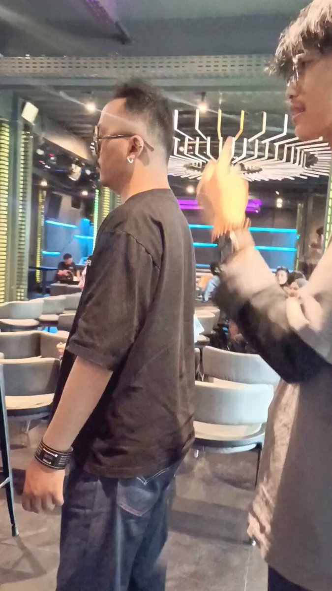 The image shows a man wearing glasses standing in front of another person who has their arm around them, possibly playing ping pong together. The background features several chairs and dining tables arranged for people to sit and eat. There are also other people present in the scene, indicating that this is likely taking place at an event or gathering where people can enjoy food and socialize.