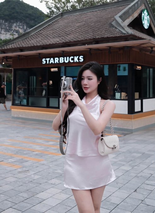 A young woman in a white sleeveless top and skirt takes a selfie with her phone outside a Starbucks coffee shop with a distinctive traditional-style roof, while carrying a white quilted handbag.
