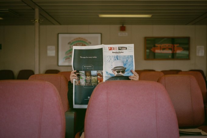 A person reads a Sunday paper featuring an 'Icon SundayStyles' section in a room with pink seats, partially obscuring their face.